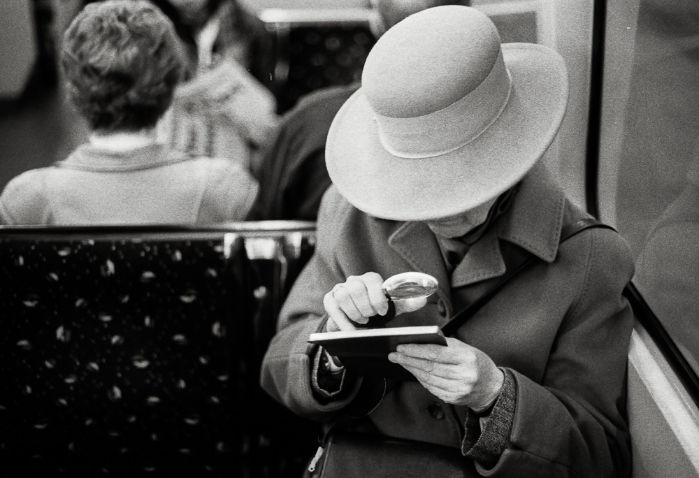 A black and white photo of an older woman wearing a wide-brimmed hat and a winter coat, sitting on public transit. She is holding a small book and examining it closely using a magnifying glass.