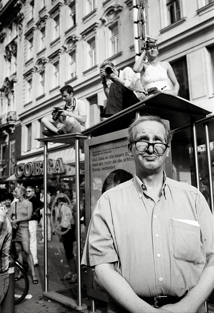 A black and white street photo featuring an older man with thick glasses in the foreground, looking into the distance with a mildly bemused expression. Behind him, two people are sitting on the roof of a glass bus shelter, taking pictures with cameras.