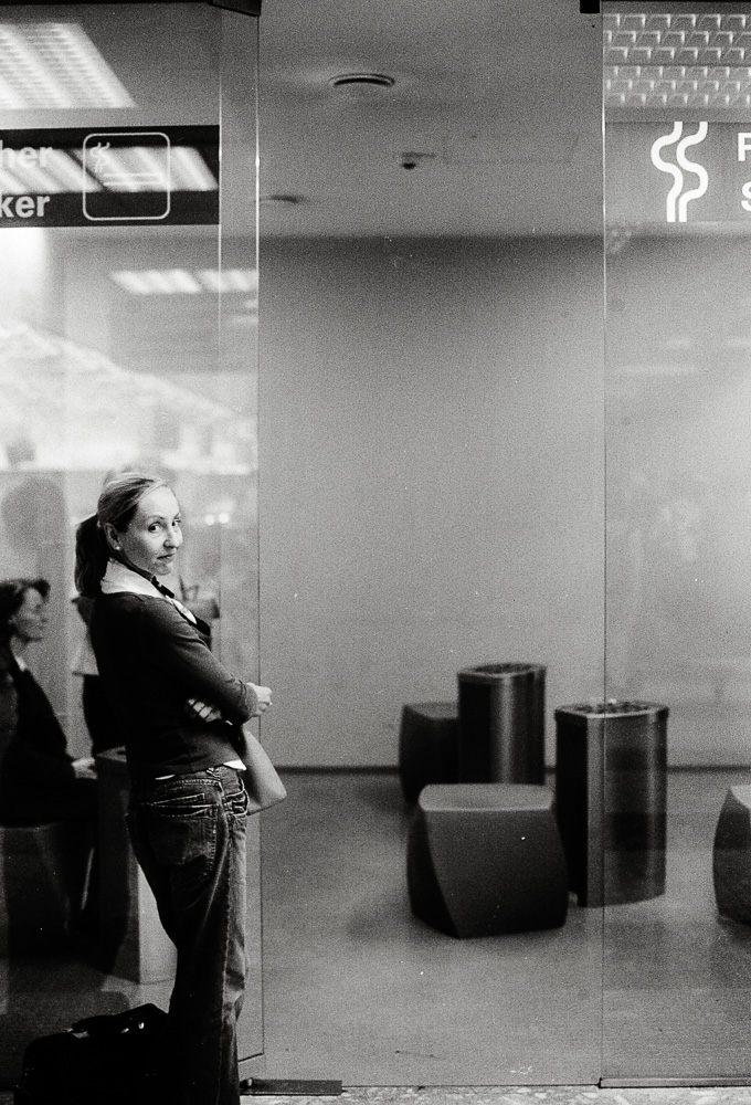A black and white photo of a woman standing beside a large glass wall in a modern indoor space. She has her arms crossed and is looking back over her shoulder directly at the camera, smiling knowingly.