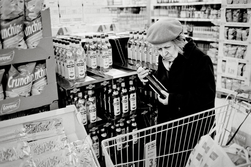 A black and white photo of an older woman wearing a beret and a dark coat in a grocery store aisle. She is holding a bottle of liquor like you would a child, ready to place it into the shopping cart parked in front of her.