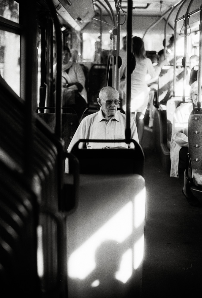 A black and white photo taken inside a bus. An older man wearing glasses and a collared shirt sits by himself looking forward. A striking patch of bright sunlight illuminates the back of the seat directly in front of him.