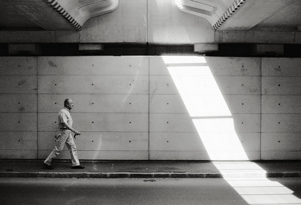 A black and white photo of a man walking along a sidewalk beneath a large concrete overpass or bridge. A sharp, bright diagonal beam of sunlight cuts across the textured concrete wall in front of him.