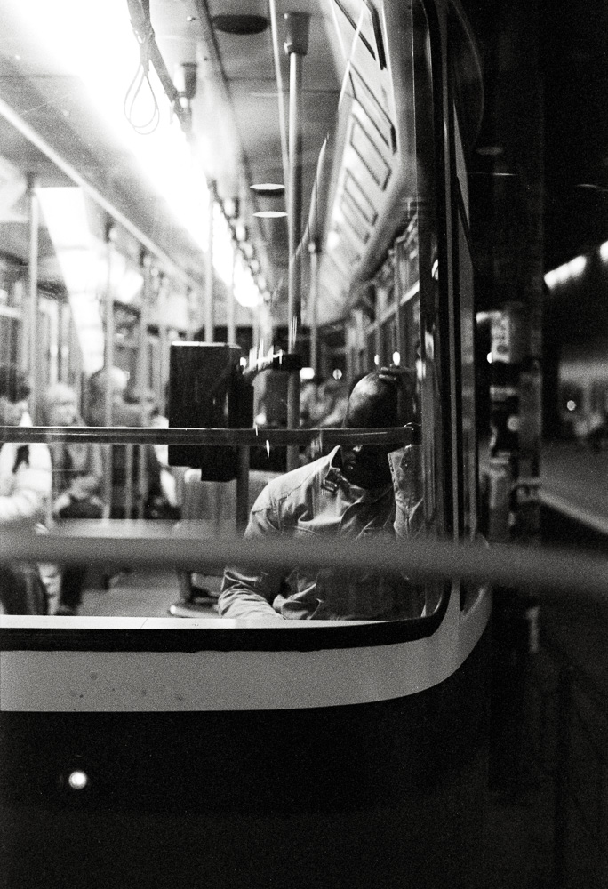 A black and white photo taken from outside a brightly lit tram at night, looking in through the window. Inside, a passenger is seated with their head resting heavily on their hand, appearing tired or lost in thought.