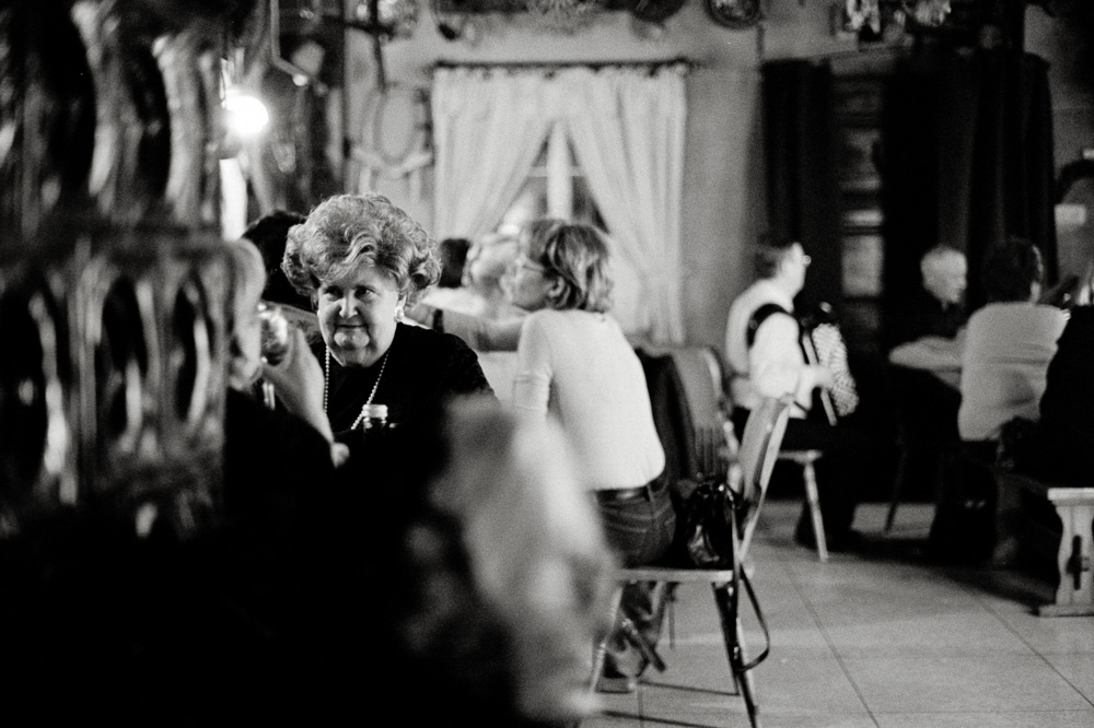 A black and white photo set inside a busy, dimly lit tavern or restaurant. In the foreground, an older woman with voluminous styled hair and a pearl necklace sits at a table. In the background, other patrons converse and a musician plays an accordion.