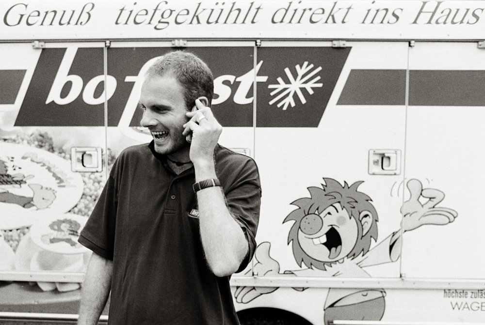 A black and white photo of a young man laughing while talking on a cell phone. He is standing in front of a "bofrost" delivery truck, which features a large, expressive cartoon character painted on the side.