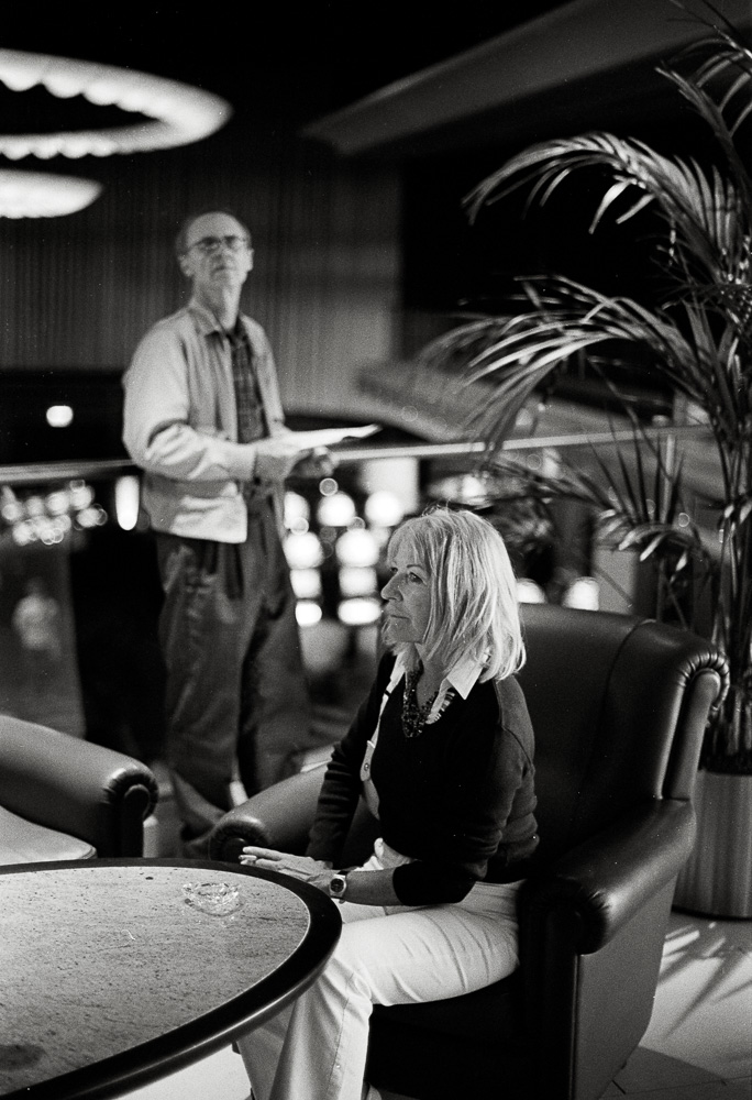 A black and white photo of two people in a lounge or hotel lobby. In the foreground, a blonde woman sits quietly in a dark leather armchair, looking off to the side. Behind her, an older man stands holding some papers and looking out over a balcony.