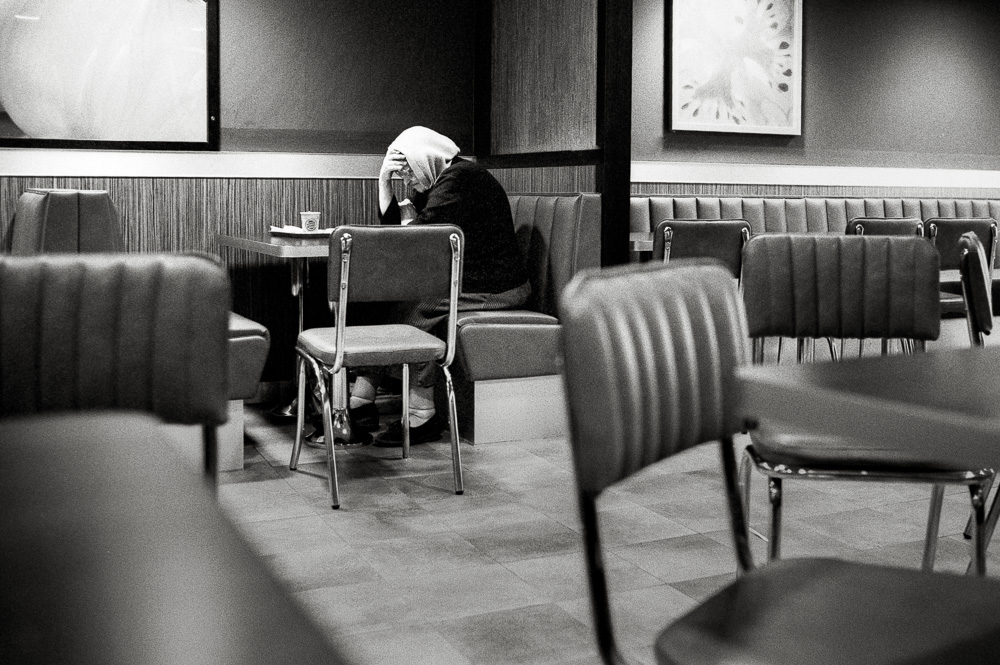 A black and white photo taken inside a fast-food restaurant. A person wearing a head covering sits alone at a table, slumped over with their head resting heavily in their hand, appearing tired or distressed.