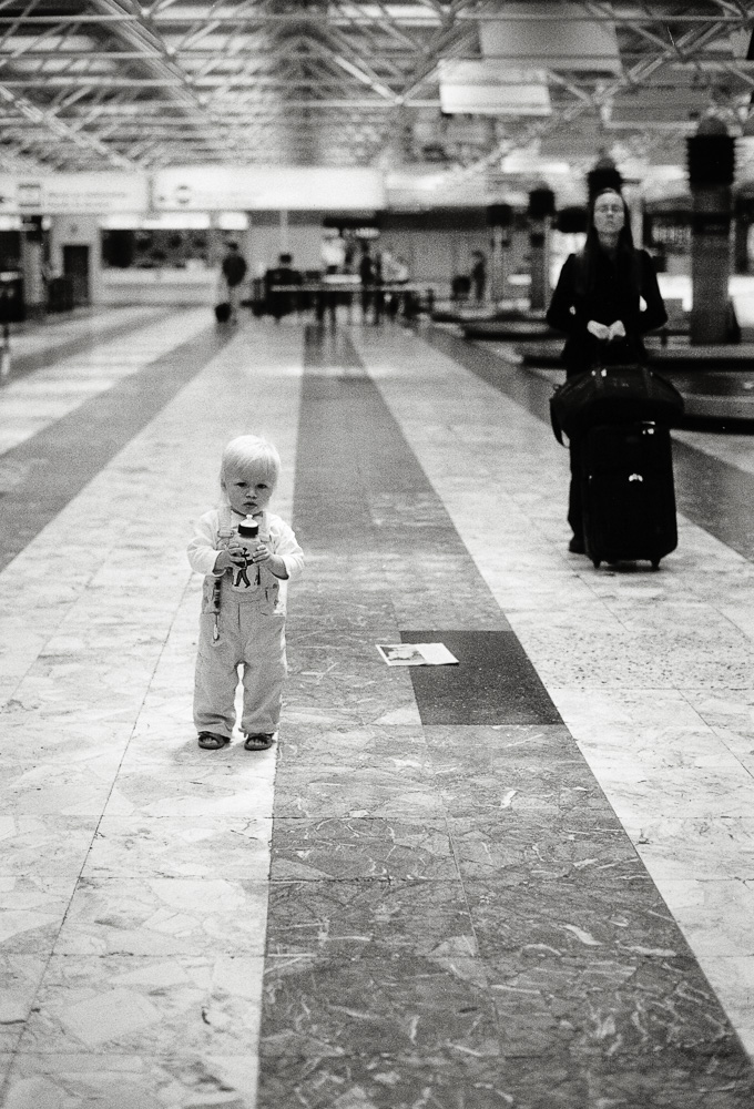 A black and white photo inside what appears to be an airport baggage claim. A toddler wearing overalls stands in the foreground holding a bottle, while an adult woman dressed in dark clothing stands behind them next to a large rolling suitcase, looking upward.