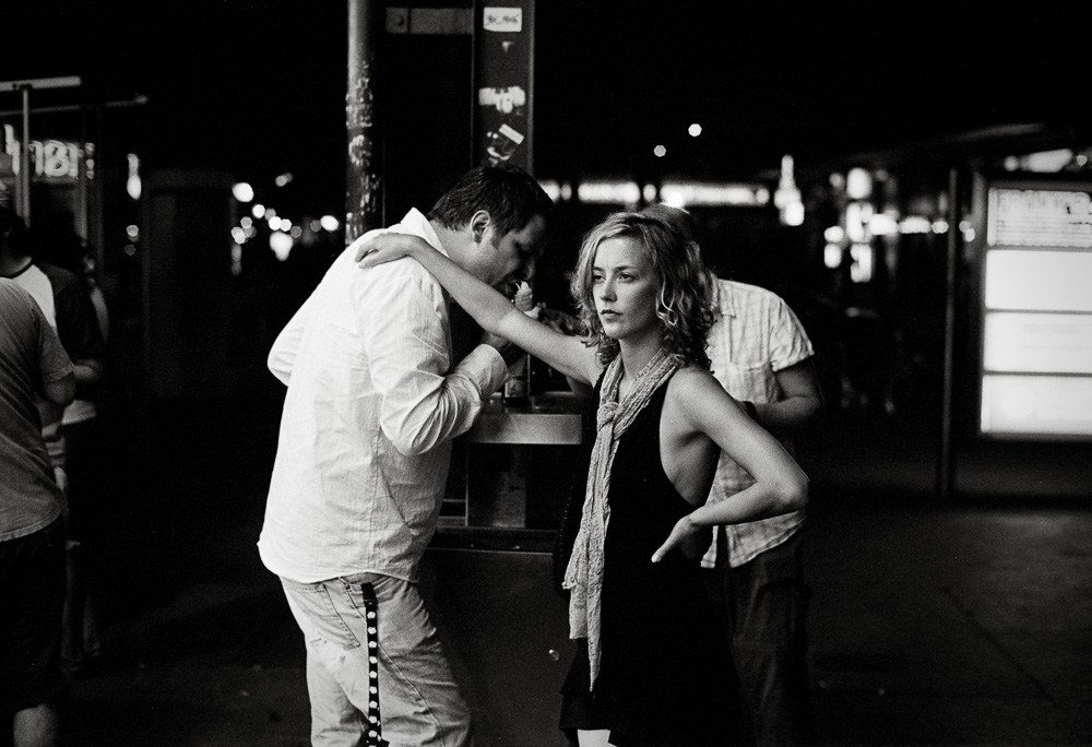 A black and white nighttime street photo. A man leans in closely toward a young woman, who is standing with her hand on her hip and looking away from him with a neutral, slightly unamused expression.
