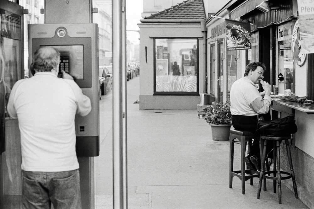 A black and white street scene. On the left, a man is standing and using an ATM. On the right, another man sits on a barstool at a small curbside food stand, eating a hot dog.
