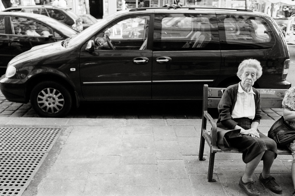 A black and white photo of an older woman with short hair sitting alone on a wooden sidewalk bench. She has a contemplative expression. A dark minivan is parked directly behind her.