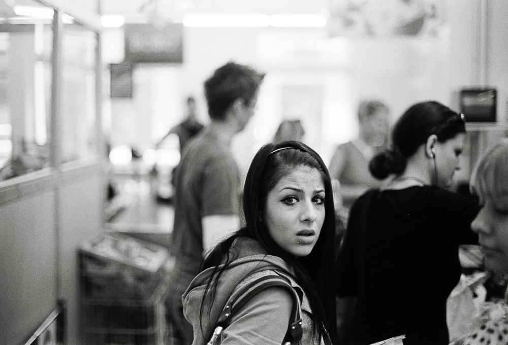 A black and white photo taken inside a busy store or checkout line. A young woman in the foreground turns to look directly at the camera with a surprised and intense expression, while other shoppers are blurred in the background.