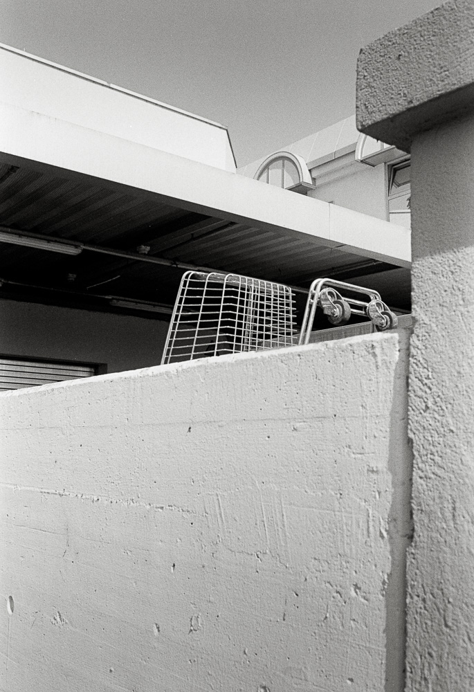 A black and white photo looking up at a thick, textured concrete wall. An empty wire shopping basket rests on top of the wall, with the flat awning and roof of a modern commercial building visible behind it.