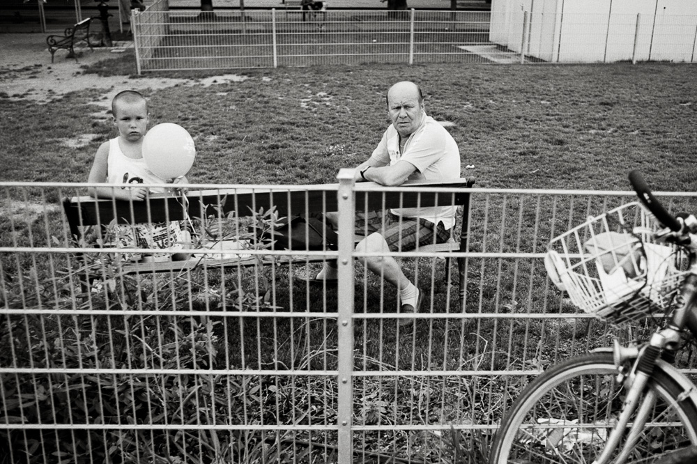 A black and white photo taken through a metal wire fence. On the other side of the fence, a young boy holding a light-colored balloon and an older man are sitting together on a bench in a grassy park. The handlebars and basket of a bicycle are visible in the right foreground.
