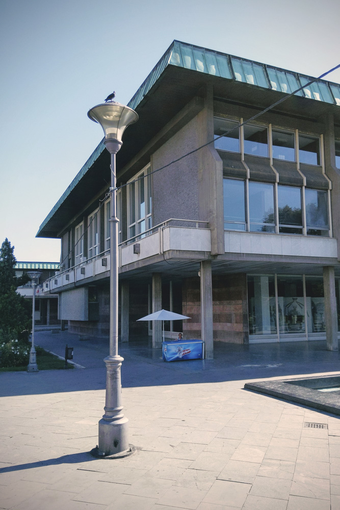A slightly faded color photo of a large concrete building with a green copper roof edge. In the foreground, a pigeon sits perched on top of a tall, classic streetlamp. On the paved plaza beneath the building, there is a small blue ice cream stand with a white umbrella.