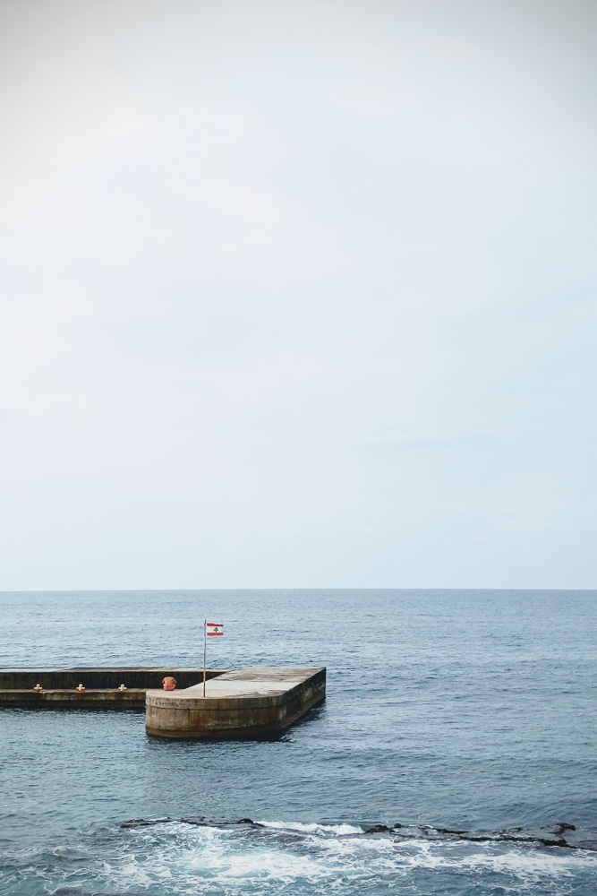 A calm, wide view of the ocean under a pale sky. A flat concrete pier extends into the water from the left. A small Lebanese flag flies on a thin pole at the edge of the pier, and a person sits quietly nearby facing the water.