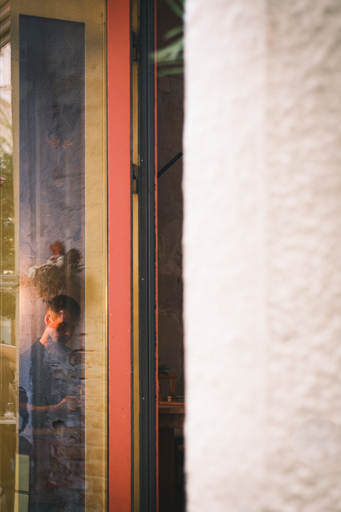 A view looking through a window with a bright red and yellow painted frame. Through the reflective glass, a person inside is visible looking down. The exterior wall next to the window has a thick, textured white surface.