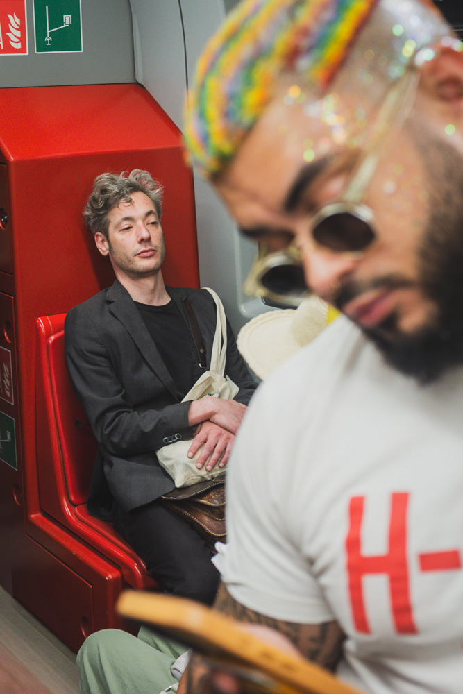 Inside a brightly lit train car. In the background, a man wearing a dark blazer sits on a bright red plastic seat, looking relaxed or tired. In the blurred foreground, a person with rainbow-colored, glittery hair and sunglasses looks down.