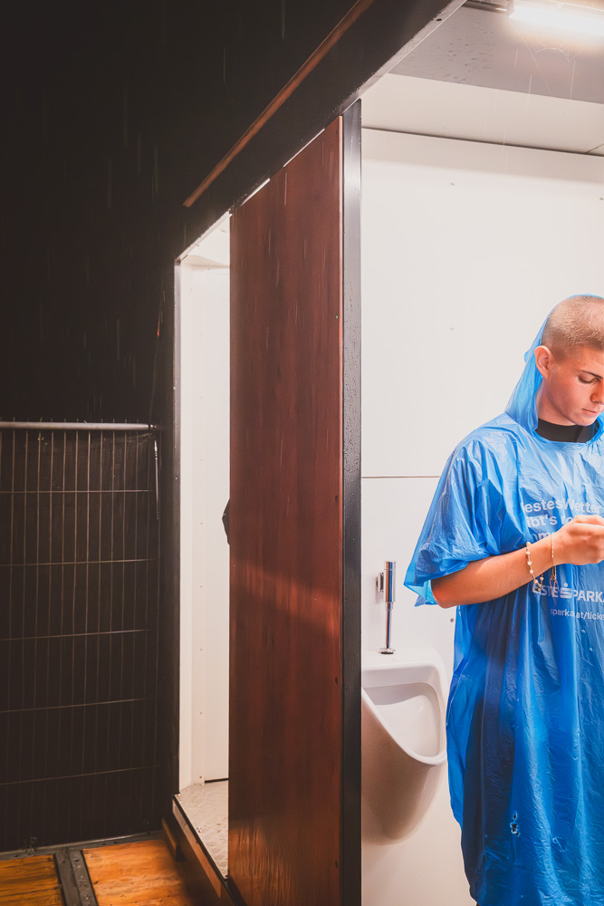 An outdoor nighttime scene showing an open portable toilet stall revealing a white urinal inside. Just outside the stall, a young person wearing a bright blue plastic rain poncho stands looking down at their phone.