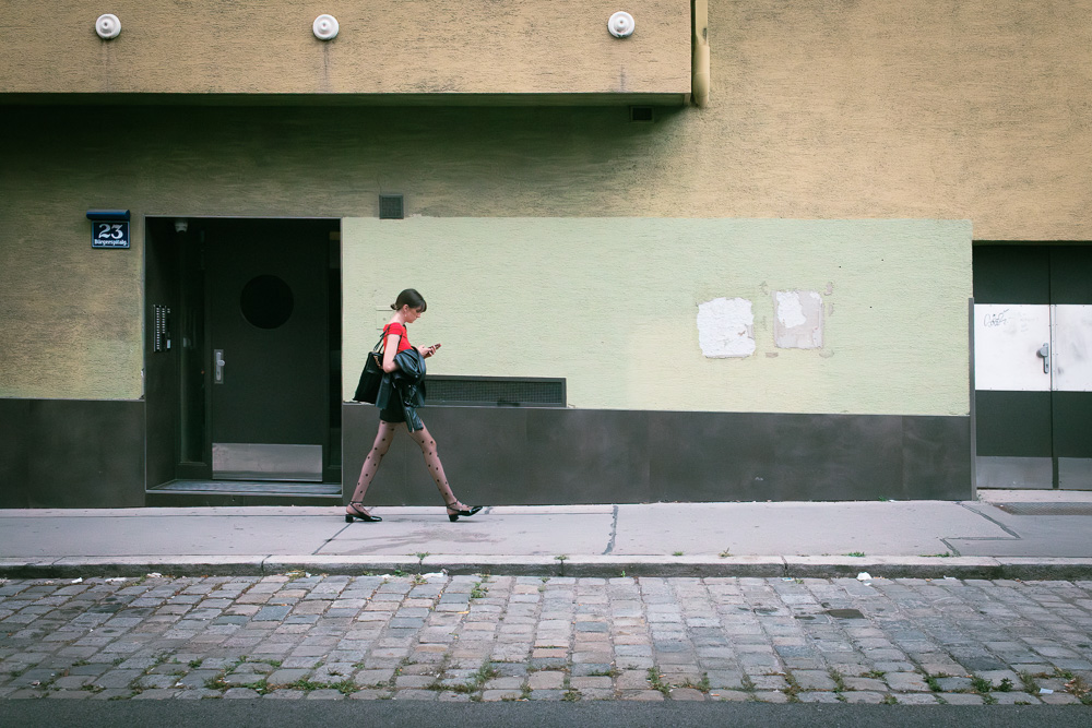 A street photograph of a woman walking briskly along a city sidewalk next to a cobblestone street. She is wearing a short red dress, a black jacket draped over her arm, and patterned black tights, and is looking at her phone. The building behind her is painted in geometric blocks of muted yellow, pale green, and brown.