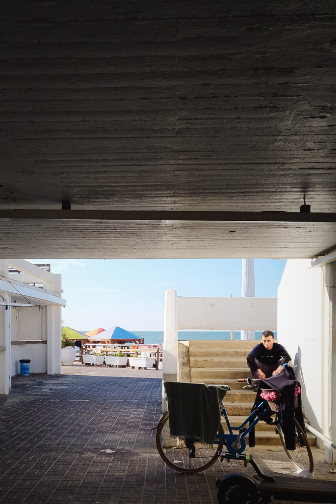 A view looking out from beneath a shaded concrete overhang toward a brightly lit beach. A man in dark clothing sits resting on a short set of white concrete stairs. In the immediate foreground, a blue bicycle and an electric scooter are parked in the shadows.