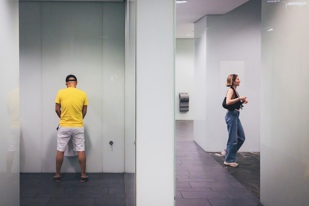 Inside a modern public restroom with white frosted glass partitions and grey tiled floors. On the left side of a glass wall, a man in a yellow t-shirt stands using a urinal. On the right side, a woman in a black top and jeans is walking through the corridor.