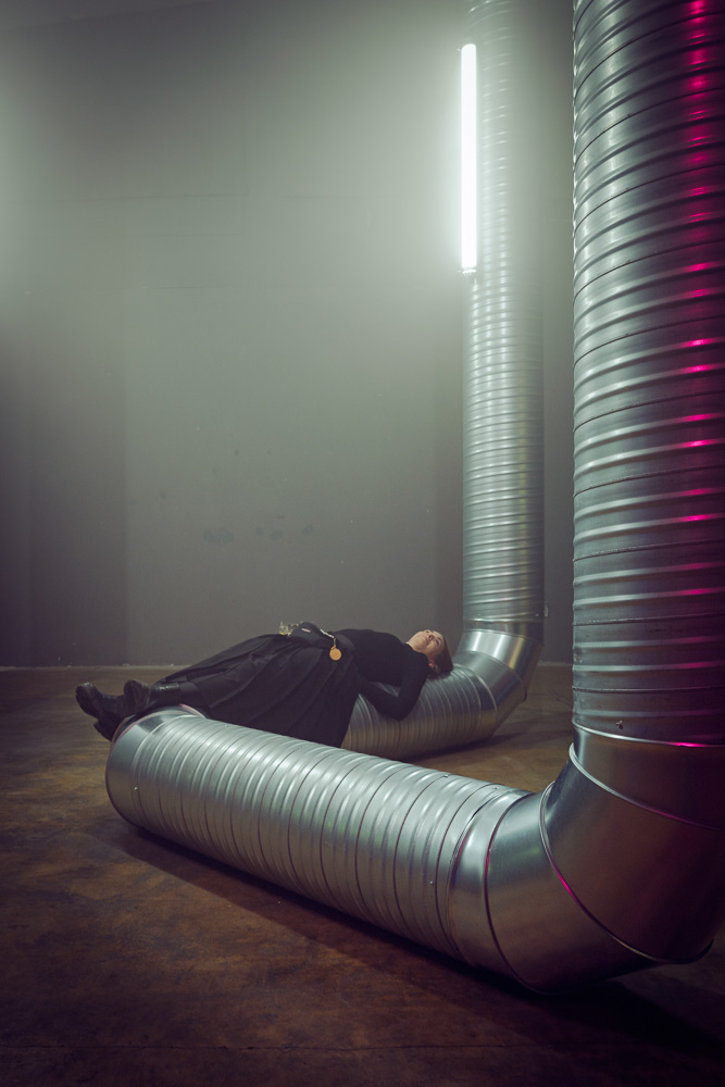 An industrial-looking room with a concrete floor and dark grey walls. A large, ribbed silver ventilation duct curves across the floor. A person dressed entirely in black is lying horizontally, resting their back directly across the metal duct beneath a bright fluorescent light.
