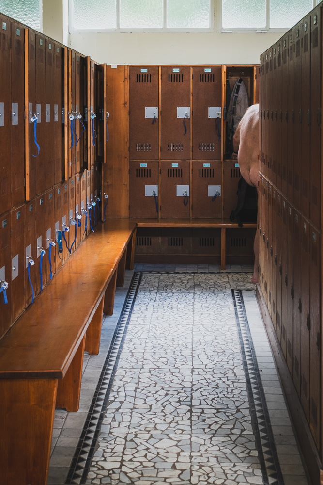 A classic locker room featuring rows of wooden lockers with blue tags hanging from their locks, and a patterned mosaic tile floor. Further down the aisle on the right, the bare back and legs of a person are partially visible as they stand at an open locker.