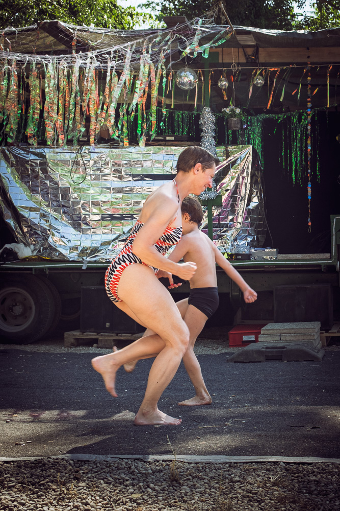 An outdoor daytime scene featuring a woman in a patterned swimsuit and a young boy in dark swim trunks running and dancing playfully barefoot on dark pavement. Behind them is a makeshift stage decorated with reflective silver foil, green tinsel, and a small disco ball.