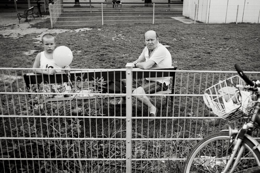 A black and white photo taken through a metal wire fence. On the other side of the fence, a young boy holding a light-colored balloon and an older man are sitting together on a bench in a grassy park. The handlebars and basket of a bicycle are visible in the right foreground.