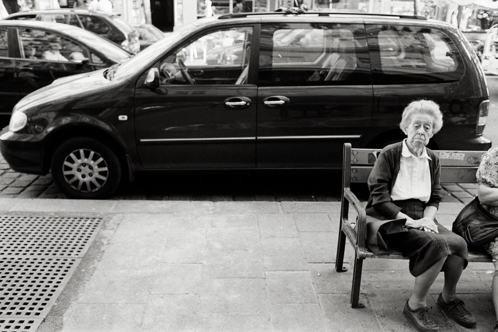 A black and white photo of an older woman with short hair sitting on a sidewalk bench, her hands folded in her lap. A second person sits beside her at the edge of the frame, and a dark minivan is parked directly behind them.