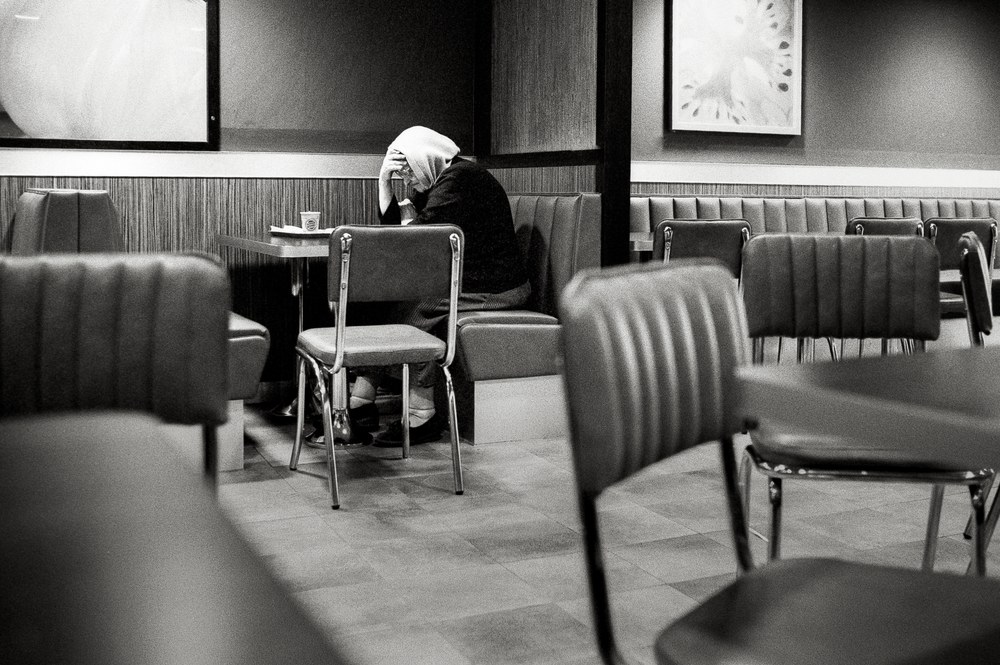 A black and white photo taken inside a fast-food restaurant. A person wearing a head covering sits alone at a table, slumped over with their head resting heavily in their hand.