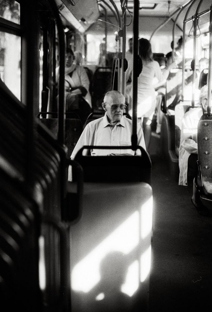 A black and white photo taken inside a bus. An older man wearing glasses and a collared shirt sits by himself looking forward. A striking patch of bright sunlight illuminates the back of the seat directly in front of him.