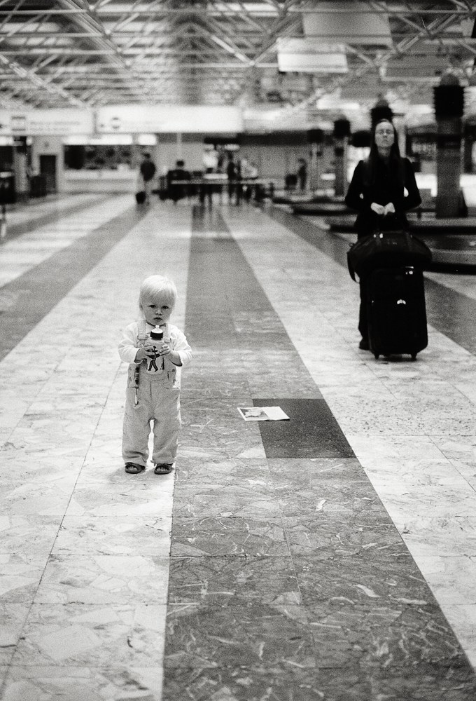 A black and white photo inside what appears to be an airport baggage claim. A toddler wearing overalls stands in the foreground holding a bottle, while an adult woman dressed in dark clothing stands behind them next to a large rolling suitcase, looking upward.