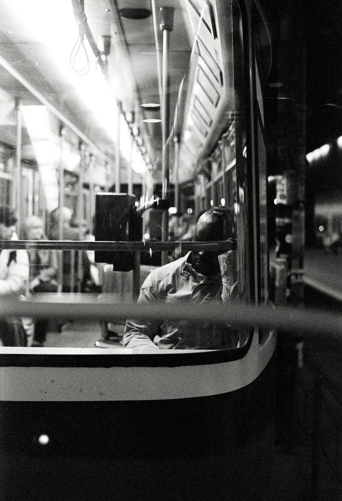 A black and white photo taken from outside a brightly lit tram at night, looking in through the window. Inside, a man sits with one hand resting on top of his head, his face mostly hidden behind a pole.
