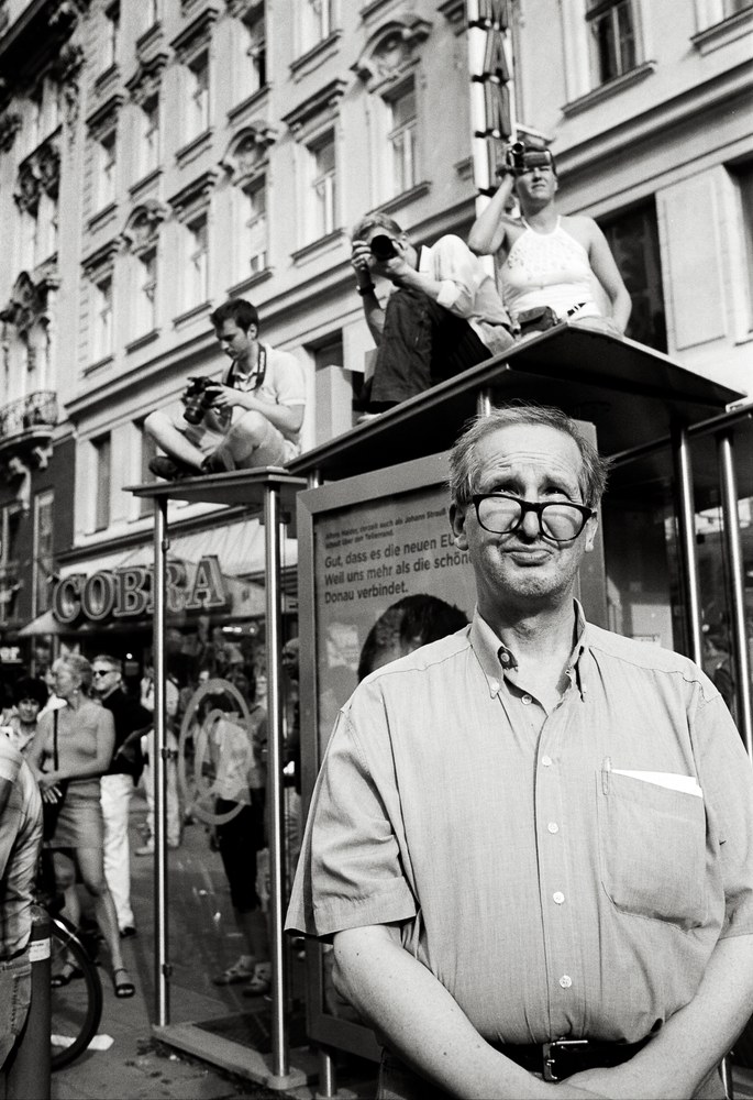 A black and white street photo featuring an older man with thick glasses in the foreground, looking into the distance. Behind him, two people are sitting on the roof of a glass bus shelter, taking pictures with cameras.
