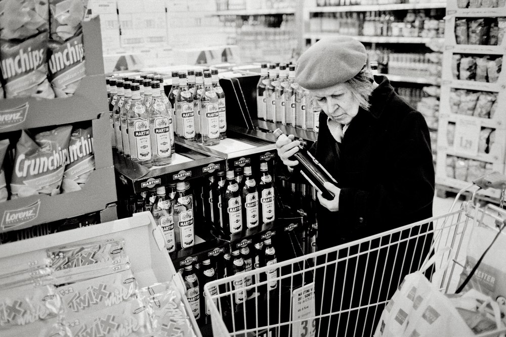 A black and white photo of an older woman wearing a beret and a dark coat in a grocery store aisle. She holds a dark bottle in both hands and looks down at it, standing behind her shopping cart next to a display of Martini bottles.