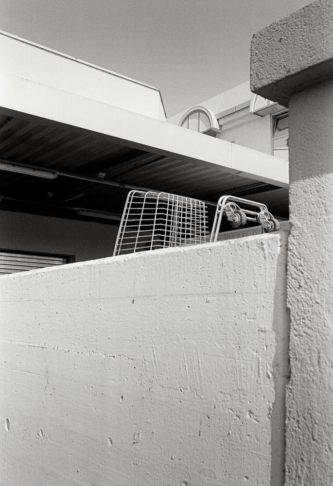 A black and white photo looking up at a textured concrete wall. Behind it, an empty wire shopping cart lies tipped on its side, wheels to the right, under the flat roof of a commercial building.