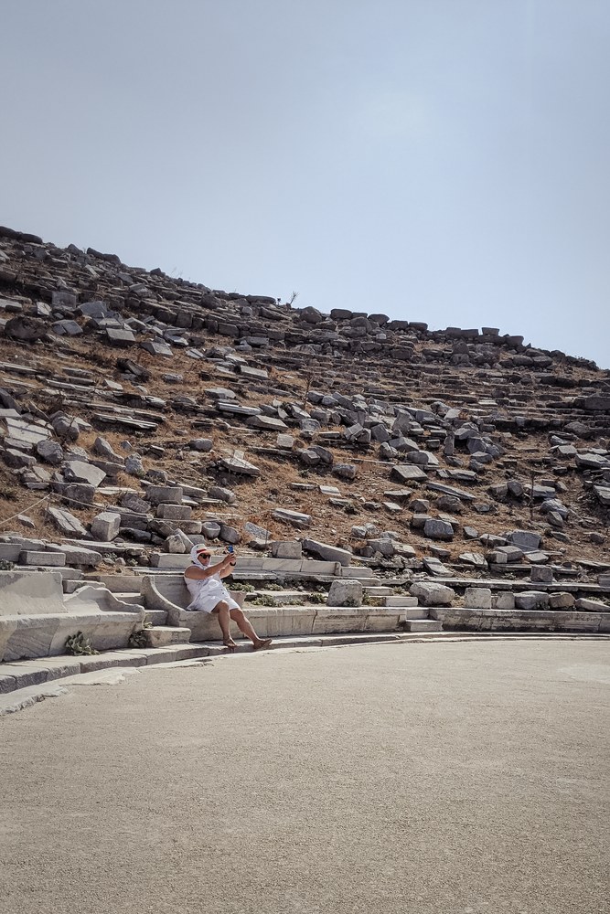 A woman in a white dress and a wide-brimmed white hat sits on the stone tiers of an ancient amphitheater, holding a phone up in front of her.