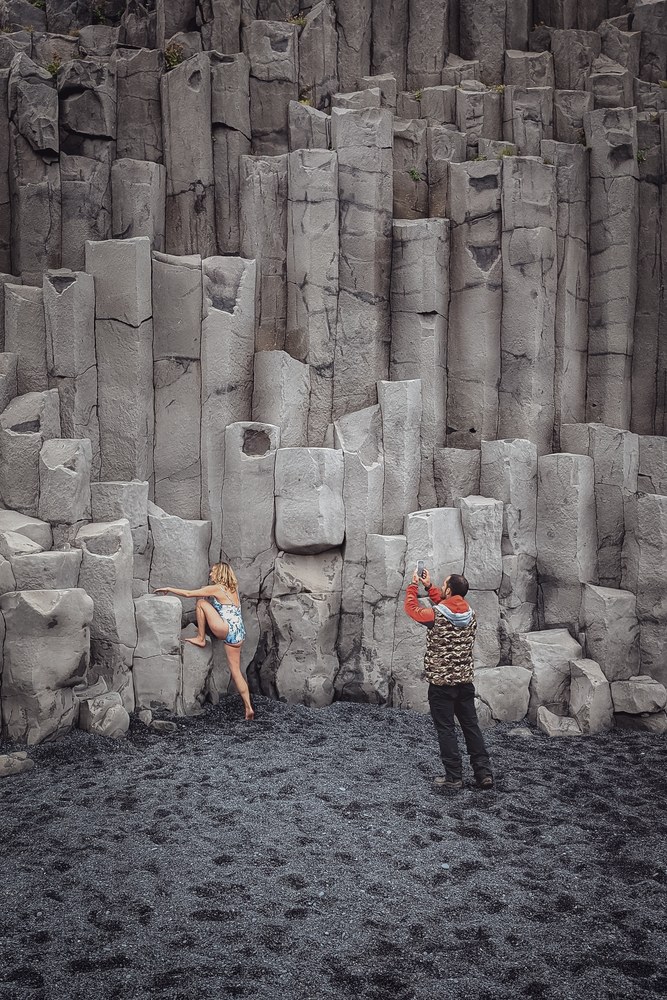 A color photo of a black sand beach with tall basalt columns. A woman in a light blue and white swimsuit poses barefoot against the rock, while a man in an orange jacket and camouflage vest photographs her with a smartphone.