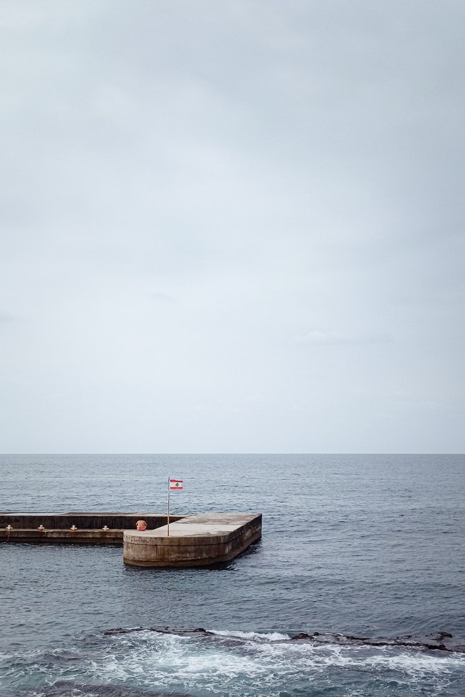 A calm, wide view of the ocean under a pale sky. A flat concrete pier extends into the water from the left. A small Lebanese flag flies on a thin pole at the edge of the pier, and a person sits nearby facing the water.