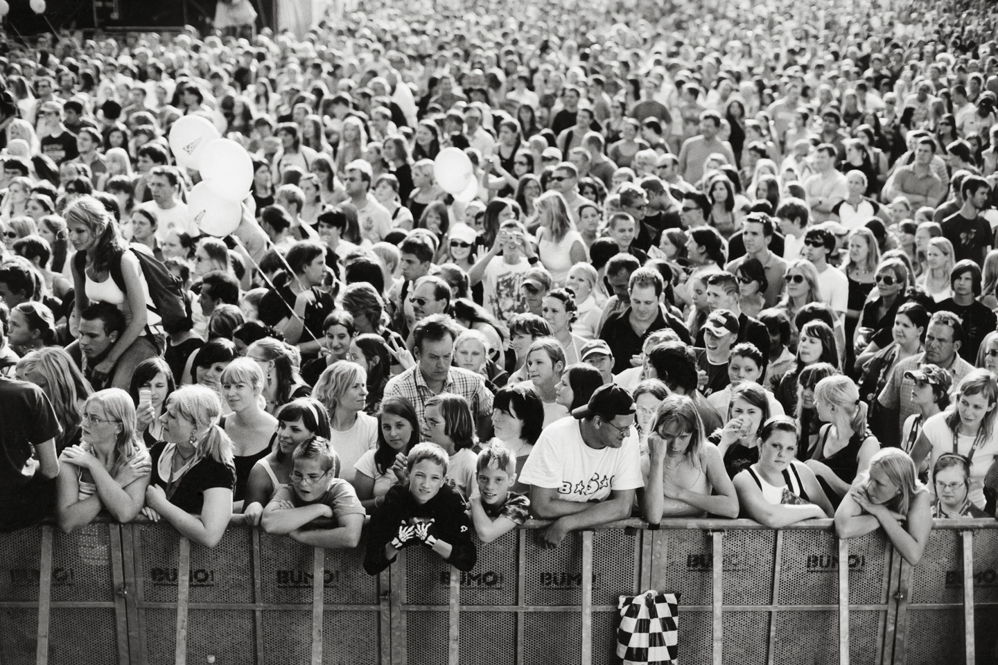 black and white photograph of a densely packed festival crowd pressed against a metal barrier. contrasting the expected excitement, the people in the front row appear exhausted and bored, leaning heavily on the rail. specific details emerge from the sea of unsmiling faces: a young boy resting his chin on hands wearing skeleton-print gloves, one person standing with their back completely turned to the stage, a woman sitting on someone's shoulders, and three white balloons floating above the crush.